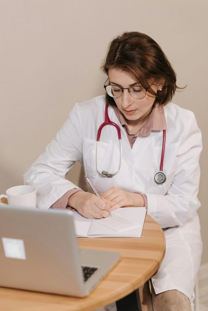 Female doctor with stethoscope writing notes in an office setting with laptop.